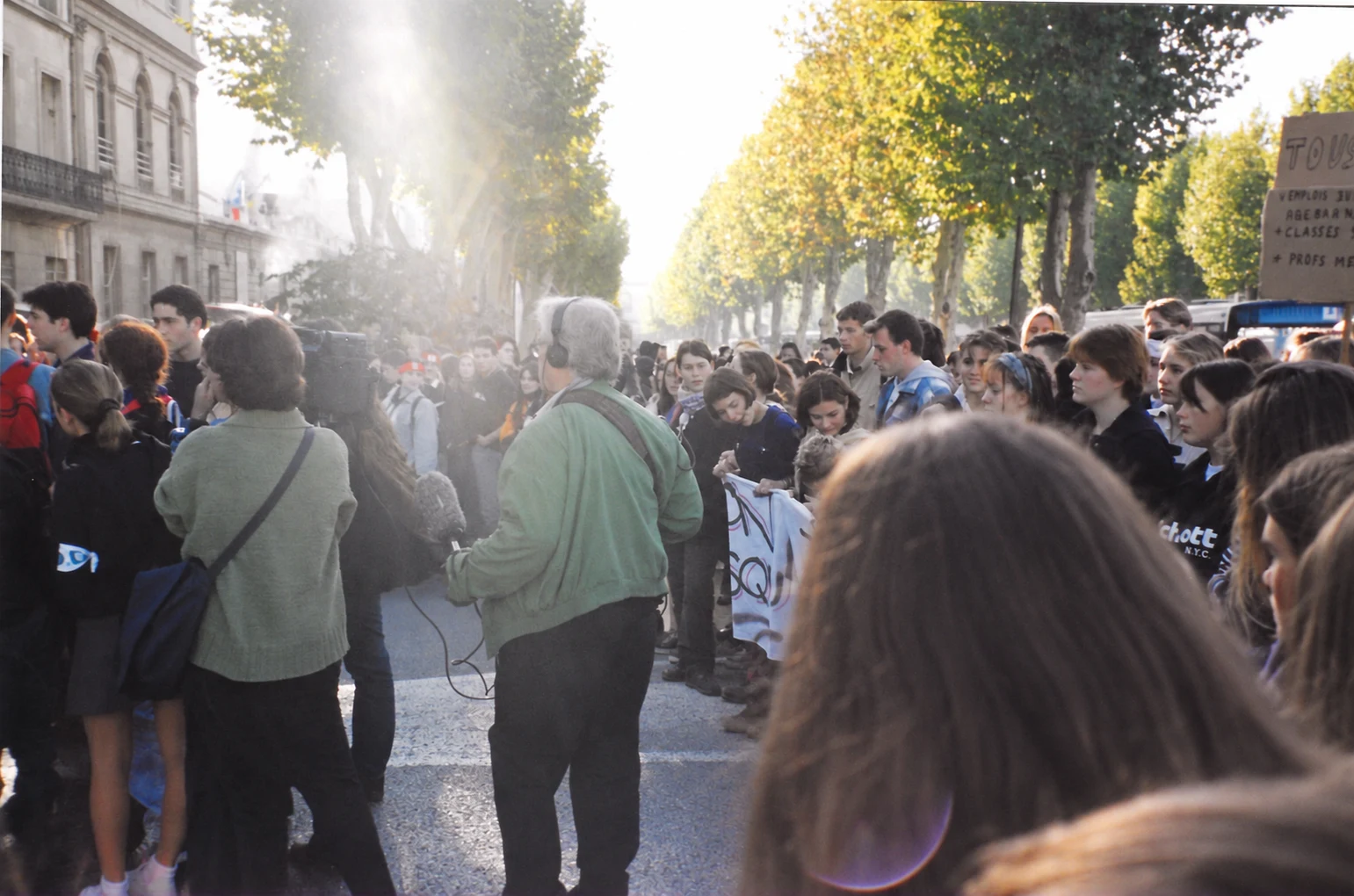 A camera operator and reporter among student demonstrators on a tree-lined street in Nîmes.