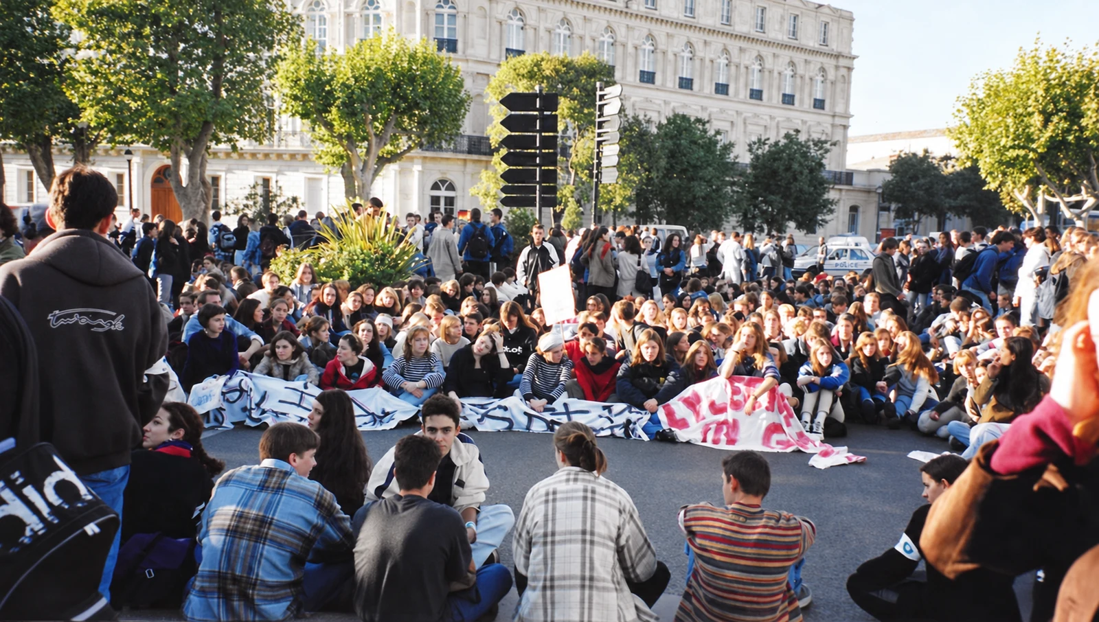 Students sitting in the street behind protest banners, with more demonstrators gathered around them.