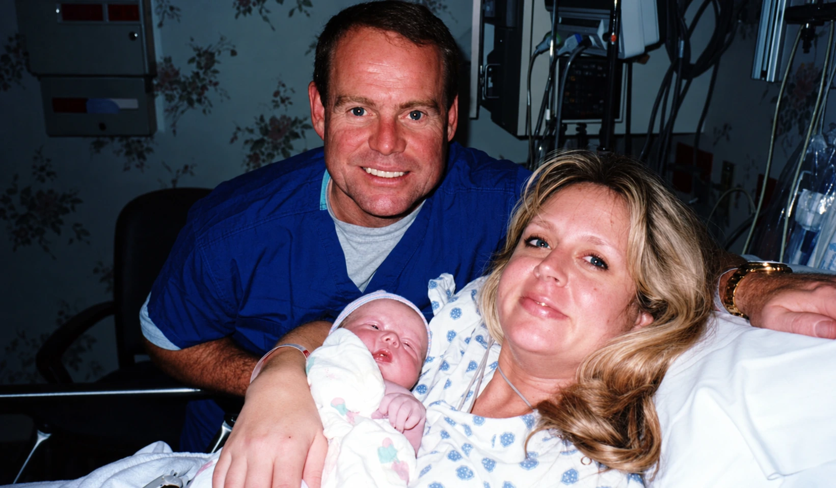 Perry in blue scrubs beside Allison holding newborn Brock in a hospital